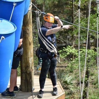 A person wearing an orange helmet and a safety harness stands on a small wooden platform on a high ropes course. They are pointing to the side while holding onto a rope. Large blue barrels are part of the obstacle nearby. The background shows trees and foliage, indicating the course is set in a forested area.