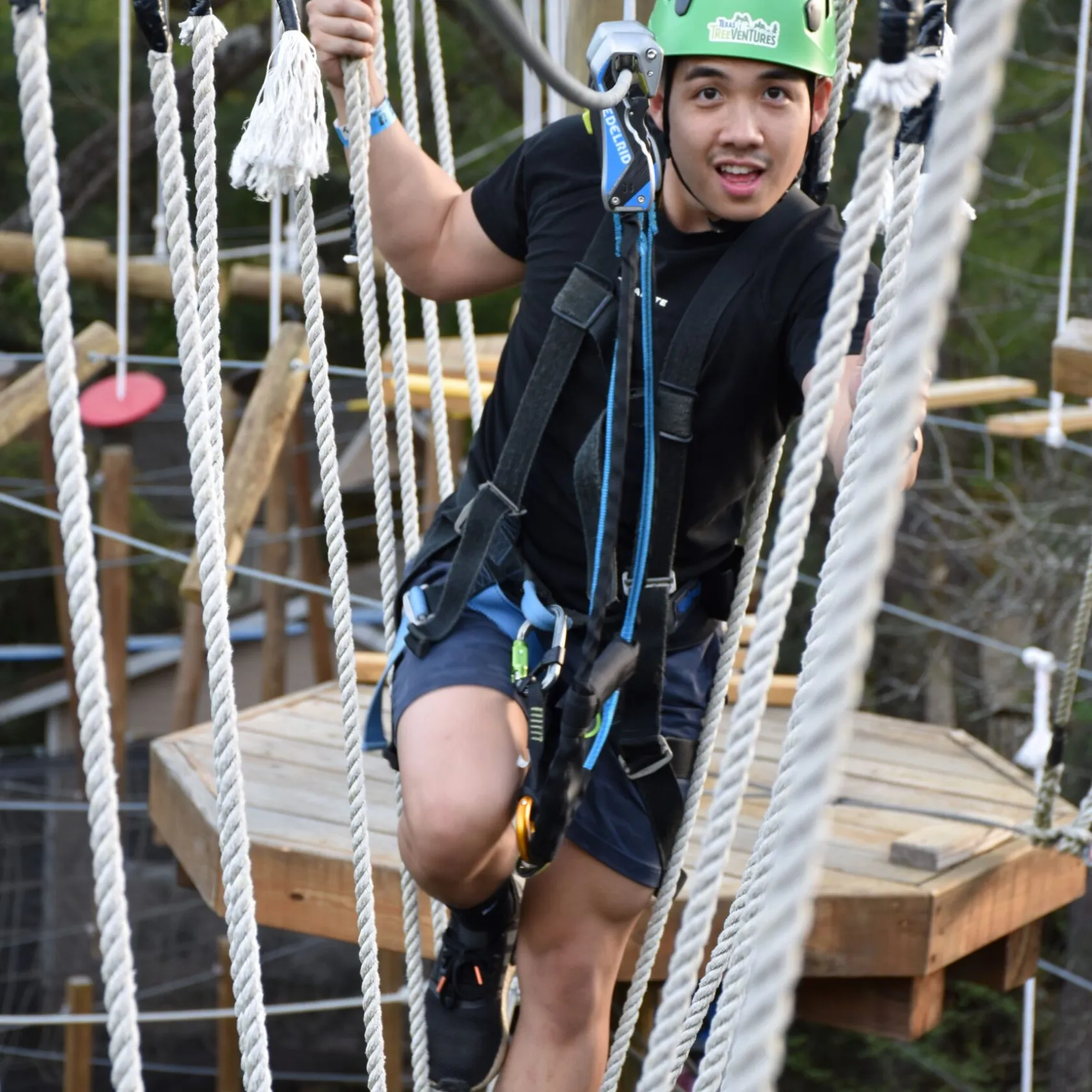 A young man wearing a green helmet and safety harness navigates a high ropes course. He is stepping between vertical ropes and holding onto them for support. The man is dressed in a black T-shirt, shorts, and athletic shoes. The background features a wooden platform and various ropes, indicating an outdoor setting.