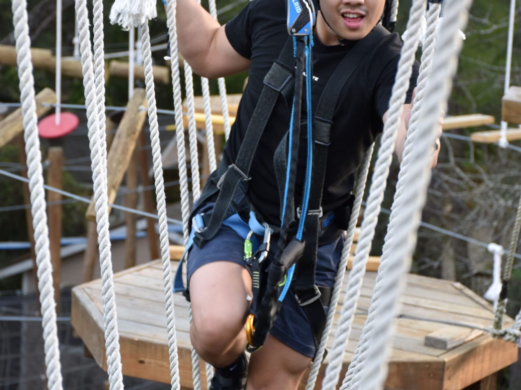 A young man wearing a green helmet and safety harness navigates a high ropes course. He is stepping between vertical ropes and holding onto them for support. The man is dressed in a black T-shirt, shorts, and athletic shoes. The background features a wooden platform and various ropes, indicating an outdoor setting.