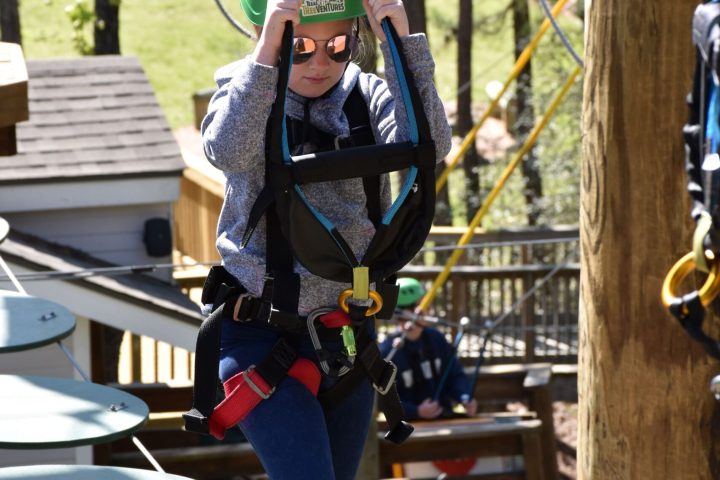 A young child wearing a green helmet and safety harness navigates a high ropes course. They are stepping on round, disc-like platforms suspended by ropes and holding onto higher ropes for balance. The child is dressed in a light blue sweater, blue pants, and sneakers, and is also wearing sunglasses. The background includes trees, a building, and other course elements, indicating an outdoor setting.