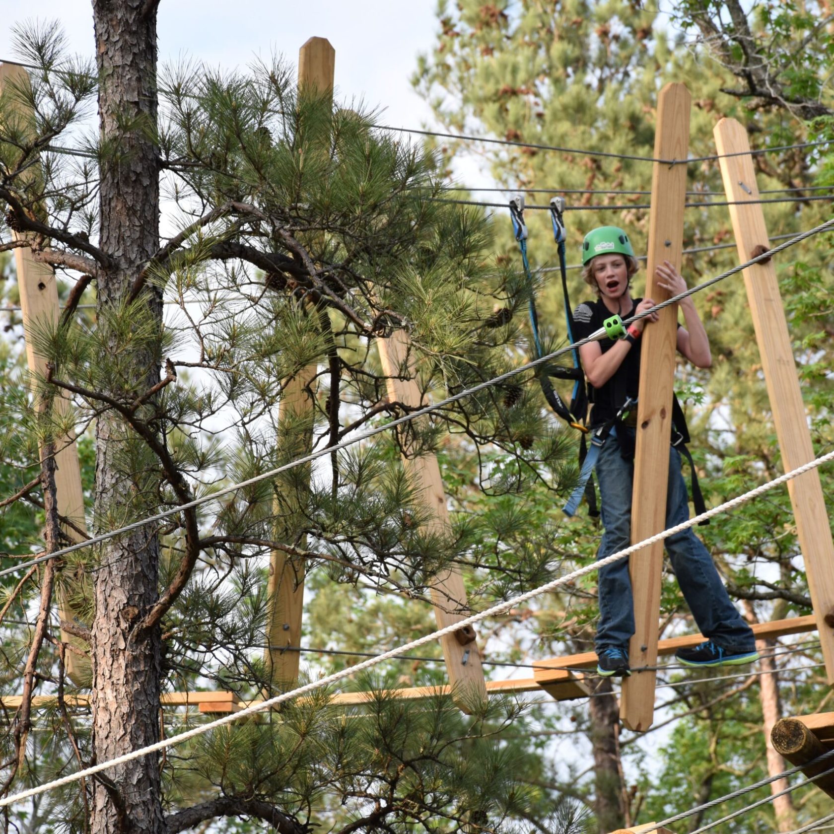 A young person wearing a green helmet and safety harness navigates a high ropes course. They are holding onto and stepping between tall wooden beams suspended in the air. The background features trees with green foliage, indicating an outdoor setting.