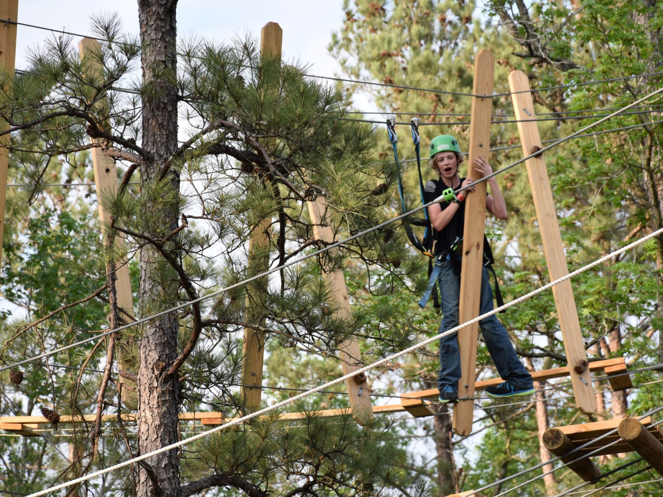 A young person wearing a green helmet and safety harness navigates a high ropes course. They are holding onto and stepping between tall wooden beams suspended in the air. The background features trees with green foliage, indicating an outdoor setting.