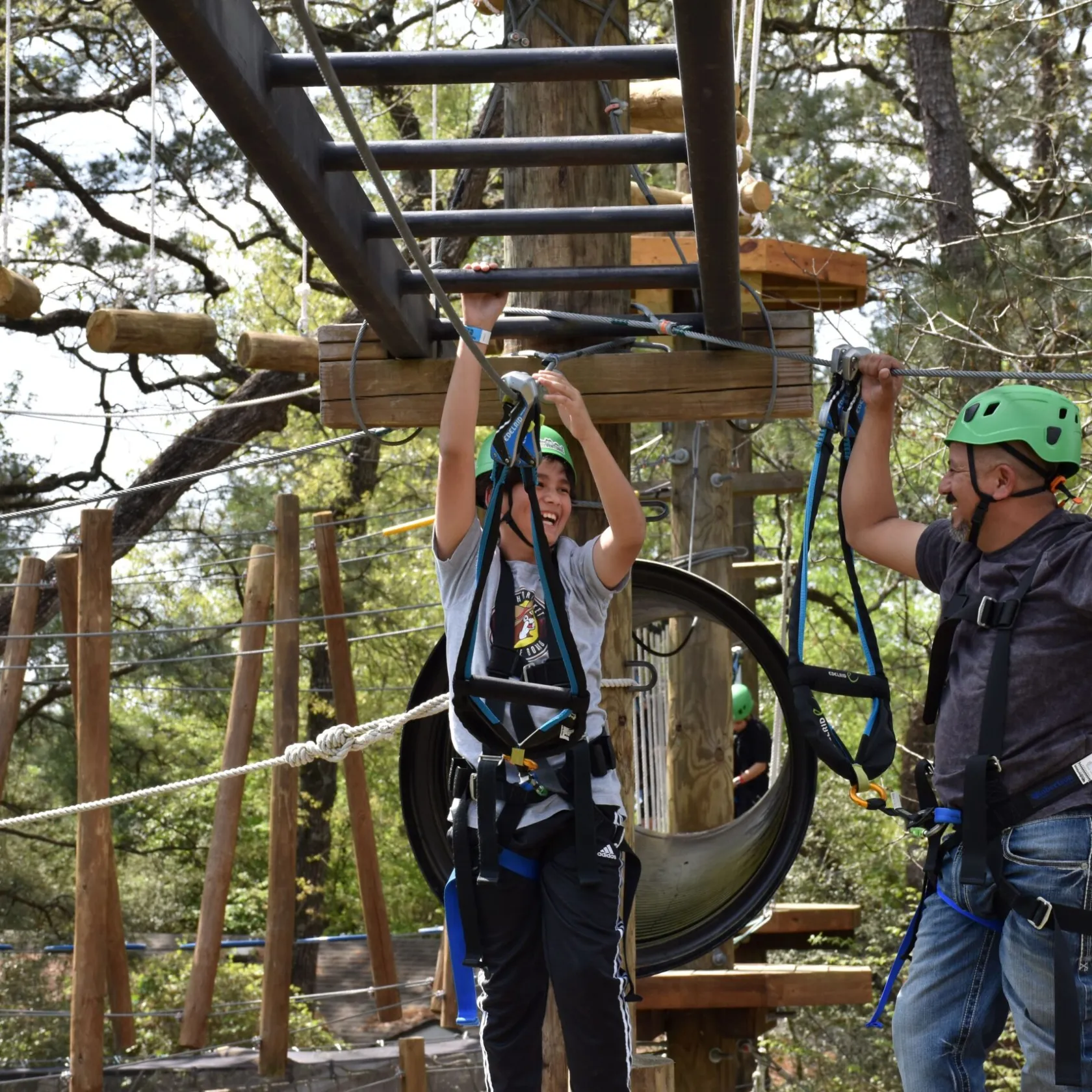 Two people wearing green helmets and safety harnesses navigate a high ropes course. One person is reaching up to grab a horizontal ladder-like structure, while the other person is holding onto a rope for balance. They are standing in front of a large vertical tube structure, and various course elements, including wooden beams and ropes, are visible in the background. The setting includes trees and foliage, indicating an outdoor environment.