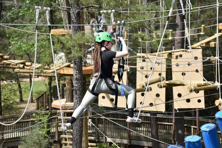 A young woman wearing a green helmet and safety harness navigates a high ropes course. She is stepping on narrow wooden planks suspended by ropes and holding onto higher ropes for balance. The woman is dressed in a black shirt, shiny silver leggings, and white sneakers. The background features various course elements, including a vertical obstacle with circular holes and blue barrels, as well as trees and foliage, indicating an outdoor setting.