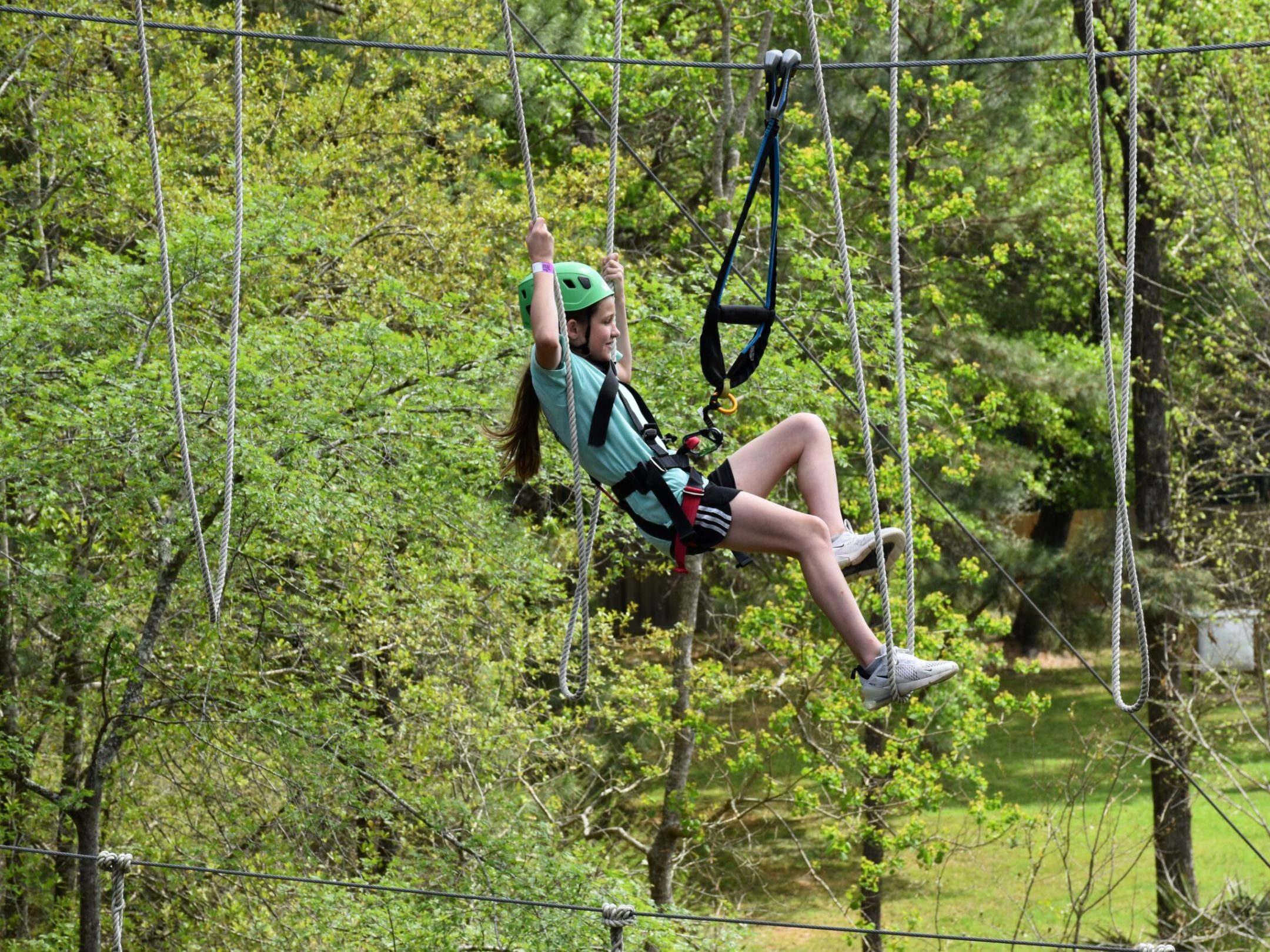 A young girl wearing a green helmet and safety harness is suspended in the air on a high ropes course. She is holding onto ropes and appears to be enjoying the experience. The girl is dressed in a light blue T-shirt, black shorts, and sneakers. The background features lush green foliage, indicating an outdoor setting.