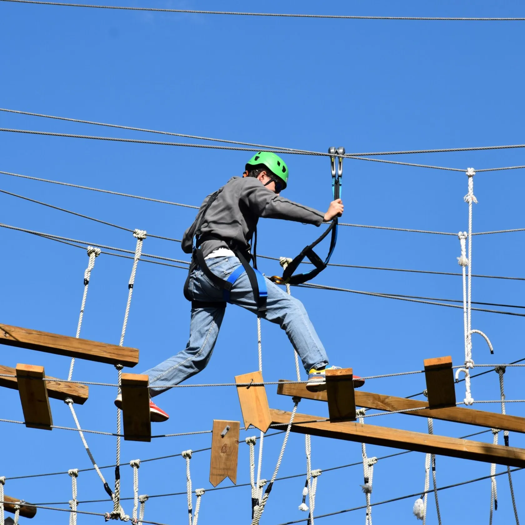 A person wearing a green helmet and safety harness navigates a high ropes course. They are stepping on narrow wooden planks suspended by ropes and holding onto higher ropes for balance. The person is dressed in a gray jacket, blue jeans, and athletic shoes. The background features a clear blue sky, indicating a sunny outdoor setting.