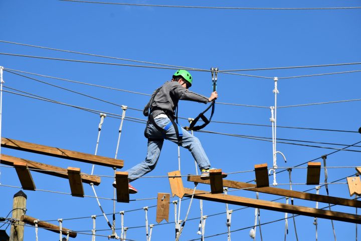 A person wearing a green helmet and safety harness navigates a high ropes course. They are stepping on narrow wooden planks suspended by ropes and holding onto higher ropes for balance. The person is dressed in a gray jacket, blue jeans, and athletic shoes. The background features a clear blue sky, indicating a sunny outdoor setting.