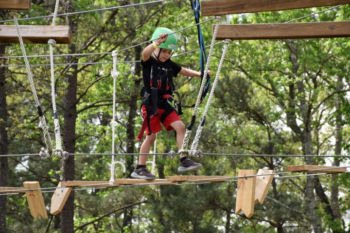 A young child wearing a green helmet and safety harness navigates a high ropes course. They are stepping on narrow wooden planks suspended by ropes and holding onto higher ropes for balance. The child is dressed in a black T-shirt, red shorts, and athletic shoes. The background features lush green foliage, indicating an outdoor setting.