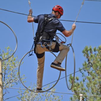 A man wearing a red helmet and safety harness navigates a high ropes course. He is stepping through large metal rings suspended in the air and holding onto ropes for balance. The man is dressed in a dark shirt and khaki pants. The background features a clear blue sky and some trees, indicating an outdoor setting.